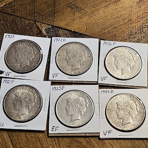 May include: Six silver Peace Dollar coins from different years, 1922D, 1925P, 1926P, 1934D, 1935, and 1922P, are displayed on white card stock with black lettering indicating the year and mint mark. The coins are graded as VF, EF, and U.