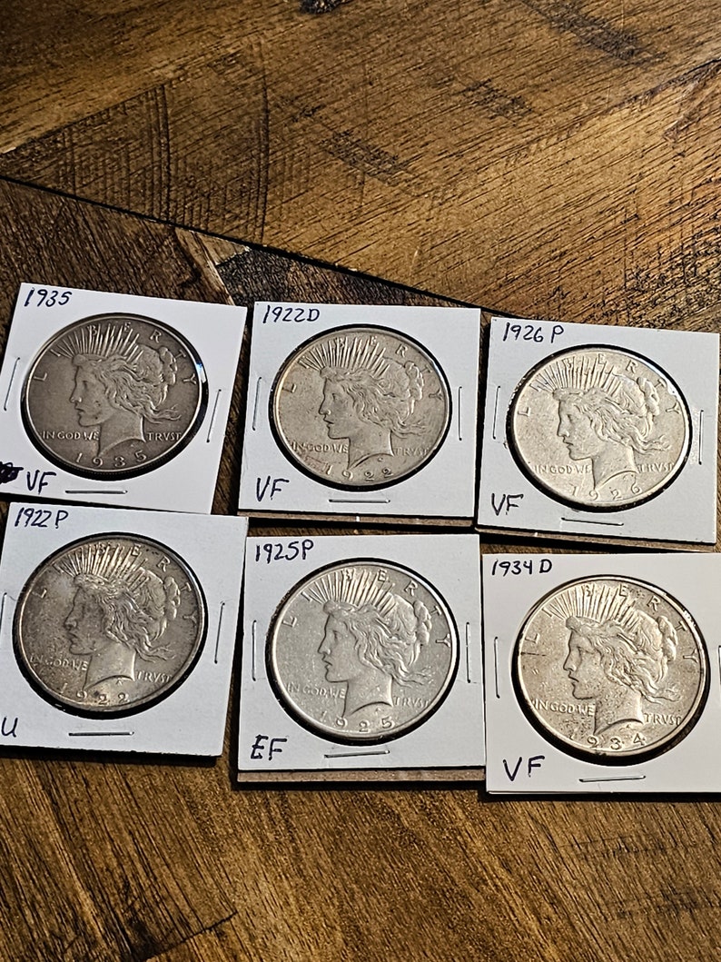 May include: Six silver Peace Dollar coins from different years, 1922D, 1925P, 1926P, 1934D, 1935, and 1922P, are displayed on white card stock with black lettering indicating the year and mint mark. The coins are graded as VF, EF, and U.