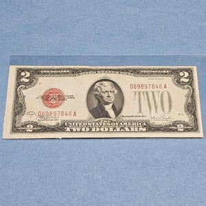 May include: A United States two-dollar bill. The bill features a portrait of Thomas Jefferson and the text "TWO DOLLARS". The bill is green and black with red seals and serial numbers. The serial number is D69897846 A.