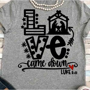 May include: A gray t-shirt with a black graphic design. The design includes the word "LOVE" with stars and polka dots, a nativity scene, and the phrase "came down" with a red heart and the text "LUKE 2:11".