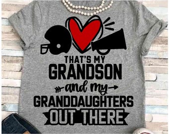 Op de afbeelding: Grijze t-shirt met de tekst "THAT'S MY GRANDSON and my GRANDDAUGHTERS OUT THERE". Het ontwerp bevat een zwarte voetbalhelm, een rood hart en een zwarte megafoon. Bovenop een witte kralenketting.