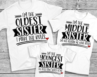 May include: Three white t-shirts with black text that reads "I'M THE OLDEST SISTER I MAKE THE RULES", "I'M THE MIDDLE SISTER I'M THE REASON WE HAVE RULES", and "I'M THE YOUNGEST SISTER THE RULES DON'T APPLY TO ME". Each shirt has a red heart.