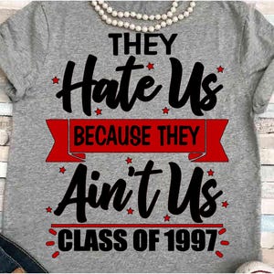 May include: A gray t-shirt with the text "THEY Hate Us BECAUSE THEY Ain't Us CLASS OF 1997" in black and red. The shirt has a crew neck and short sleeves. A pearl necklace is at the top.