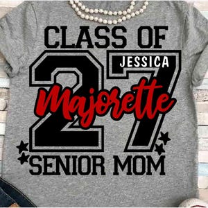 May include: A heather gray t-shirt featuring a black and red graphic design with the text CLASS OF 27, JESSICA, Majorette, and SENIOR MOM. The design includes large black numbers 27, red cursive lettering, and small black star accents.