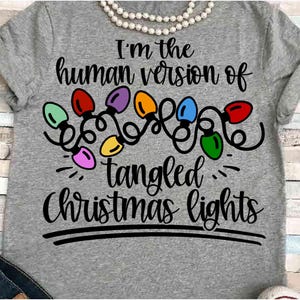 May include: A gray t-shirt with the text "I'm the human version of tangled Christmas lights." The design includes a string of colorful Christmas lights. A white pearl necklace is draped across the top.