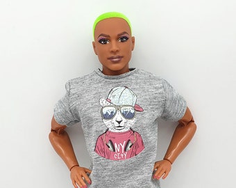 May include: A gray t-shirt with a graphic of a cartoon animal wearing sunglasses and a hat. The text "MY CITY" is printed on the shirt. The doll is wearing blue and green patterned shorts.