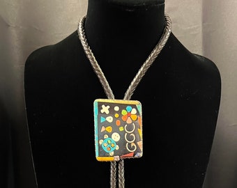 May include: A silver bolo tie with a black, square pendant featuring a colorful geometric design. The pendant is made of inlaid stones and has a silver border.