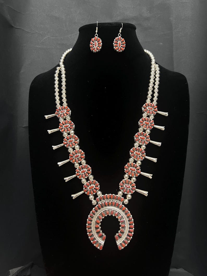 May include: A silver necklace with a horseshoe pendant and multiple round charms, all decorated with red coral stones. Matching earrings with red coral stones are also included.