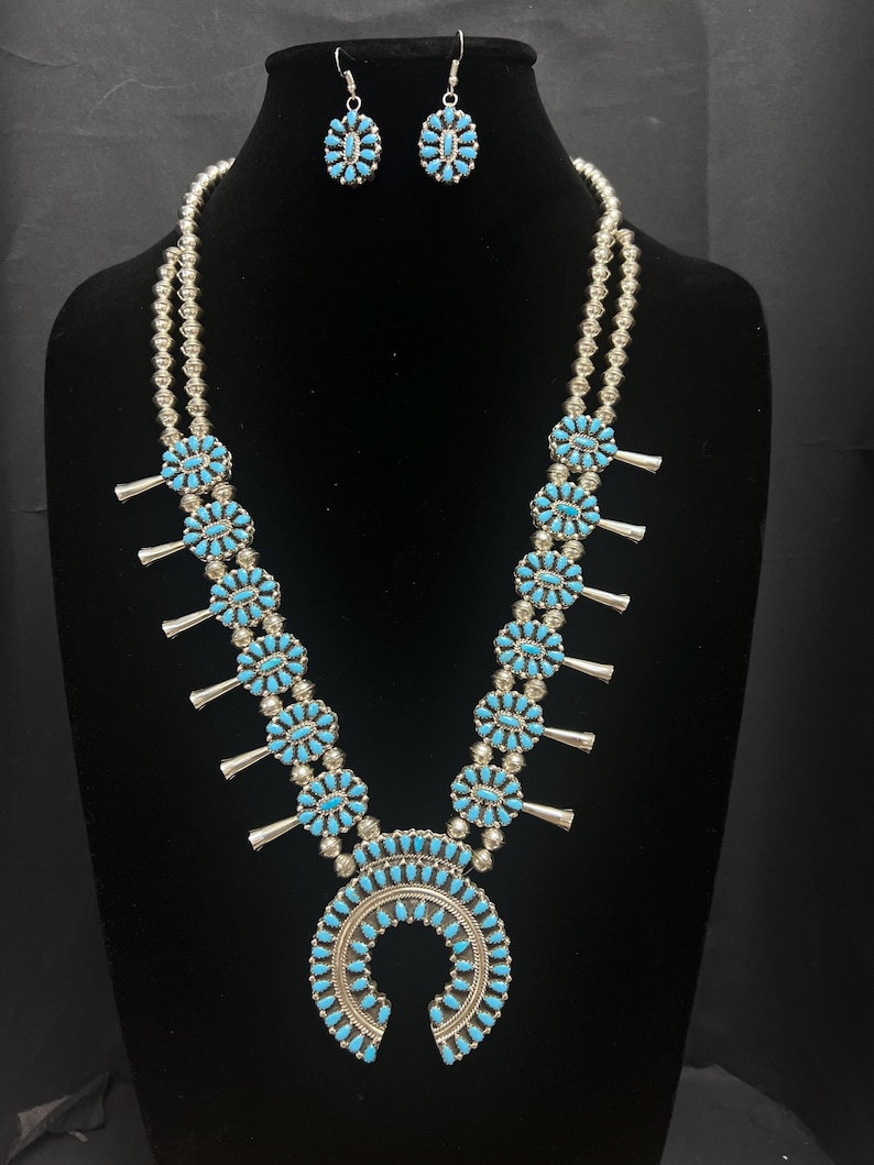 May include: A turquoise and silver Native American style necklace and earrings set. The necklace features a horseshoe pendant with turquoise stones and silver beads. The earrings are oval shaped with turquoise stones.