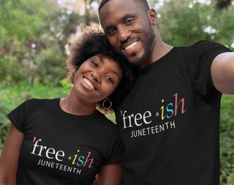 May include: Two people wearing black t-shirts with the text "free-ish" and "Juneteenth" in white and colorful lettering. The image is a close-up shot of the two people smiling, with a blurred green background.