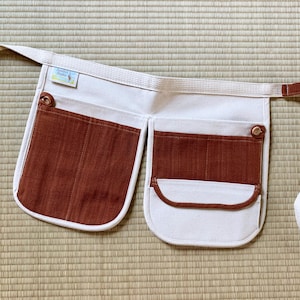 May include: A white canvas apron with brown corduroy pockets and a brown strap. The apron has a small label that says "Handmade with Love".