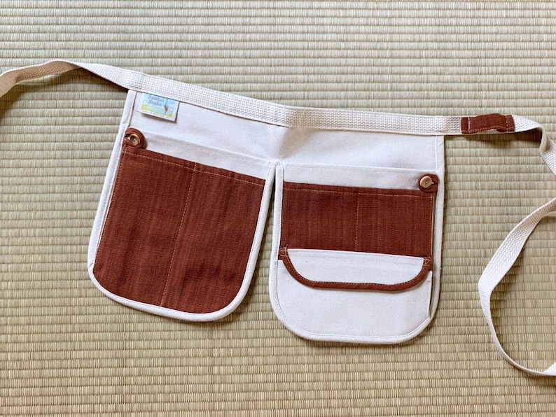 May include: A white canvas apron with brown corduroy pockets and a brown strap. The apron has a small label that says "Handmade with Love".
