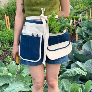 Könnte beinhalten: Eine Person trägt ein grünes Tanktop und eine Jeans-Shorts und arbeitet im Garten. Sie trägt eine weiße Baumwollschürze mit einer blauen Jeans-Tasche und einer weißen Baumwolltasche. Die Schürze hat einen weißen Gurt, der um die Taille gebunden wird. Die Person hält ein Gartenwerkzeug in der linken Hand und einen Bund grünes Blattgemüse in der rechten Hand.