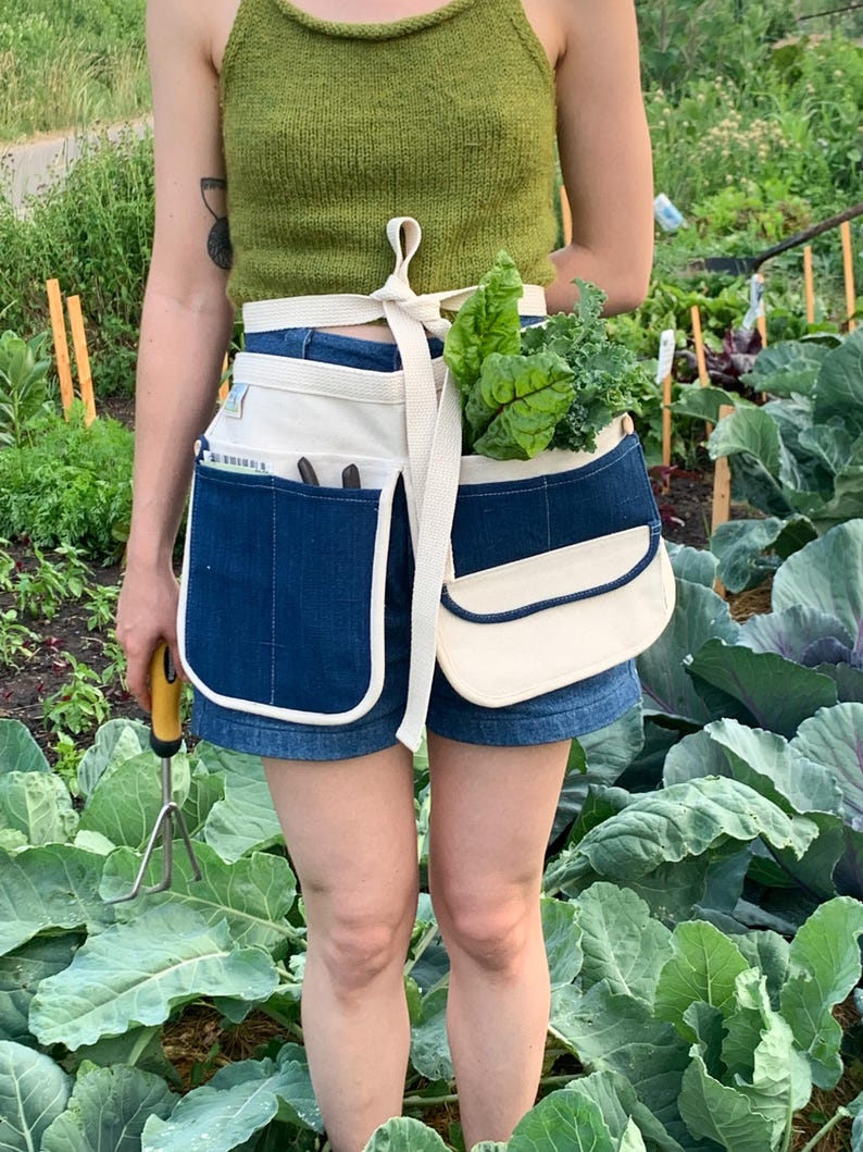 May include: A person wearing a green tank top and denim shorts is gardening. They are wearing a white canvas apron with a blue denim pocket and a white canvas pouch. The apron has a white strap that ties around the waist. The person is holding a gardening tool in their left hand and a bunch of leafy greens in their right hand.