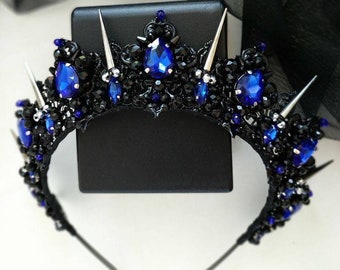May include: A black headband with silver spikes and blue rhinestones. The headband is decorated with black beads and has a crown-like shape.