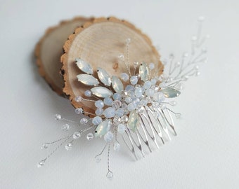 May include: A silver hair comb with a floral design featuring clear and iridescent blue crystals. The comb is displayed on a wooden surface.