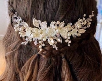 May include: A decorative hair comb featuring a silver-toned metal base adorned with clear crystals, white floral accents, and small pearl-like beads. The piece is arranged in a wide, curved shape and is secured into long, wavy brown hair.
