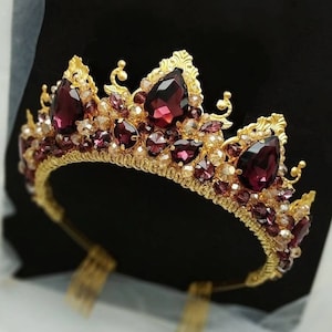 Op de afbeelding: Een goudkleurige kroon met grote, druppelvormige, dieprode edelstenen en kleinere, ronde, heldere en rode kristallen. De kroon heeft een gedetailleerd, sierlijk ontwerp en staat tegen een zwarte achtergrond.