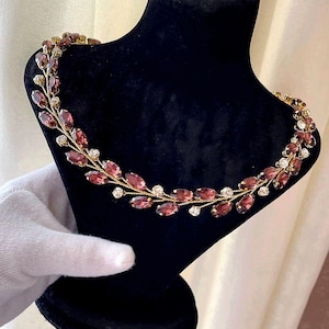 May include: A gold necklace with a delicate design featuring small, oval-shaped purple gemstones and tiny white rhinestones. The necklace is displayed on a black velvet mannequin.