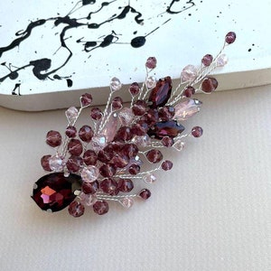 May include: A handcrafted hair accessory with purple and pink beads and crystals. The design includes a large, teardrop-shaped crystal and smaller, round beads, all attached to silver wire. This elegant piece is suitable for special occasions.
