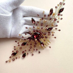May include: A gold-toned hair comb featuring a cluster of burgundy and champagne-colored beads and crystals. The comb has a central large burgundy crystal surrounded by smaller beads and crystals, creating a floral design. The comb is held by a gloved hand.