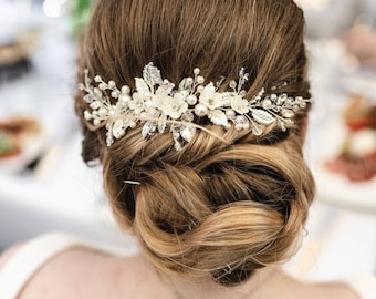 May include: A silver-toned hair comb featuring white floral accents, small round pearls, and clear crystals arranged in a botanical design with metallic leaves. The decorative piece is secured into a styled updo, showcasing intricate wire-wrapped details.