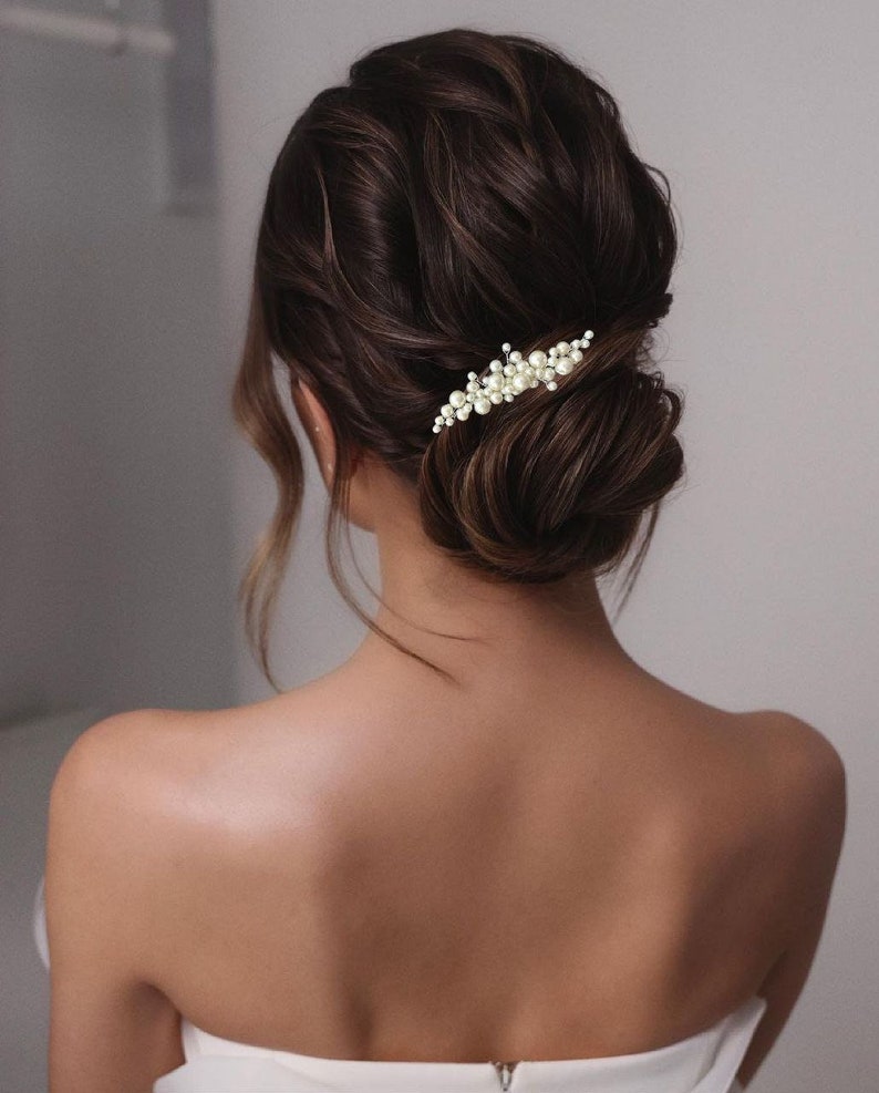 May include: A woman's updo hairstyle with a pearl hair clip. The dark brown hair is styled in a low bun with loose waves. The hair clip is a delicate design of small pearls clustered together.