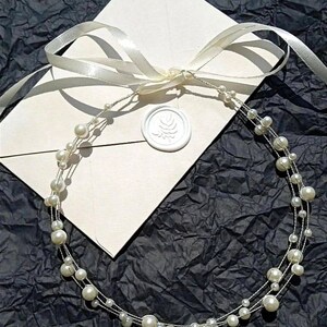 May include: A white pearl headband with multiple strands of pearls on a white envelope with a white ribbon and a wax seal.