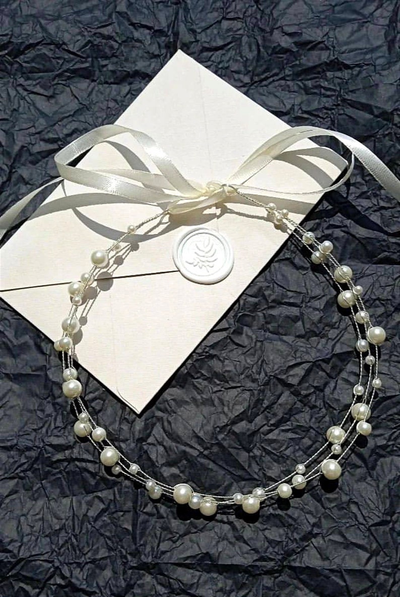 May include: A white pearl headband with multiple strands of pearls on a white envelope with a white ribbon and a wax seal.