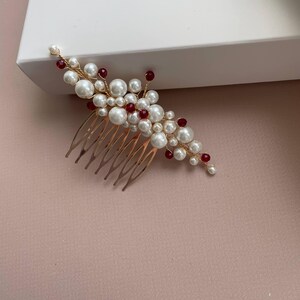 May include: A gold-toned hair comb adorned with faux pearls and red beads. The comb features a cluster of white pearls and red beads, arranged in a floral design. The comb is likely for bridal or special occasion hairstyles.