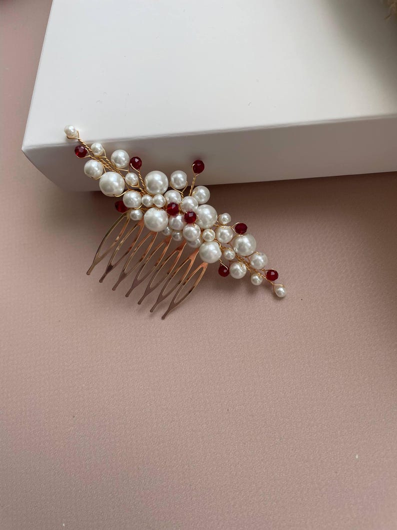 May include: A gold-toned hair comb adorned with faux pearls and red beads. The comb features a cluster of white pearls and red beads, arranged in a floral design. The comb is likely for bridal or special occasion hairstyles.