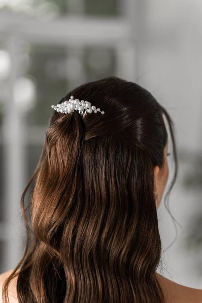 May include: A silver hair comb adorned with numerous small, round, white pearls. The comb is set in dark brown hair, styled in loose waves. The background is blurred, suggesting a focus on the hair accessory.