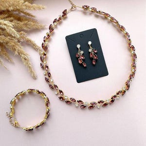May include: A set of jewellery featuring a gold necklace, bracelet, and earrings. The necklace and bracelet are adorned with purple and clear rhinestones in a leaf design. The earrings are dangle earrings with a similar leaf design.