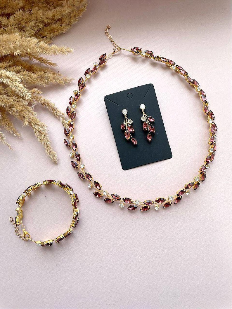 May include: A set of jewellery featuring a gold necklace, bracelet, and earrings. The necklace and bracelet are adorned with purple and clear rhinestones in a leaf design. The earrings are dangle earrings with a similar leaf design.