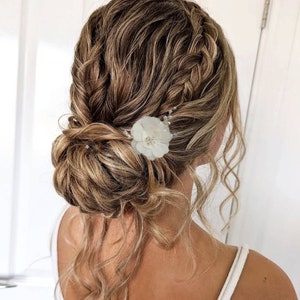 以下が含まれることがあります： 白い花のヘアピンで飾られた、編み込みのアップスタイルにした、長い明るい茶色の髪の女性。髪は緩くカールしていて、アップスタイルの周りに垂れている。