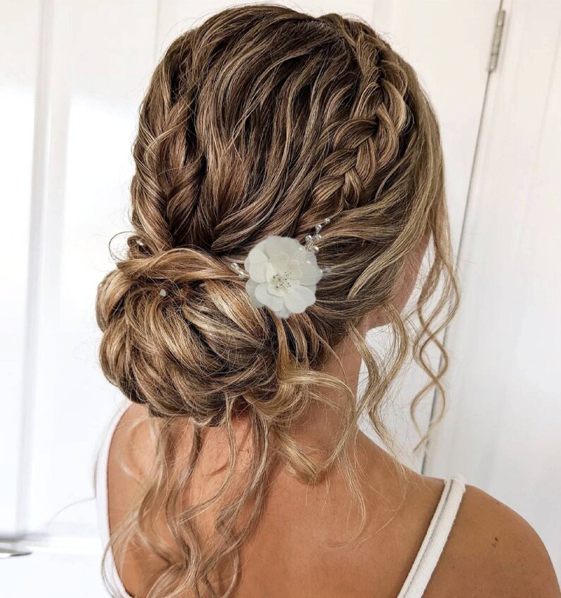 以下が含まれることがあります： 白い花のヘアピンで飾られた、編み込みのアップスタイルにした、長い明るい茶色の髪の女性。髪は緩くカールしていて、アップスタイルの周りに垂れている。