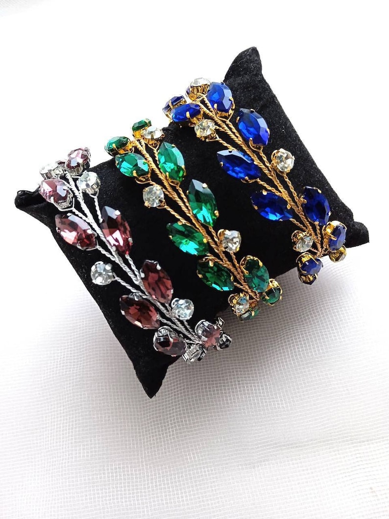 May include: Four gold leaf headbands with different coloured rhinestones. The headbands are arranged in a row on a black velvet cushion. The colours of the rhinestones are purple, green, blue, and clear.