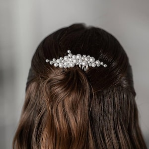 May include: A silver hair comb adorned with clusters of faux pearls. The comb is inserted into dark brown hair, styled in a half-up, half-down hairstyle. The pearls are various sizes and arranged in a floral pattern.