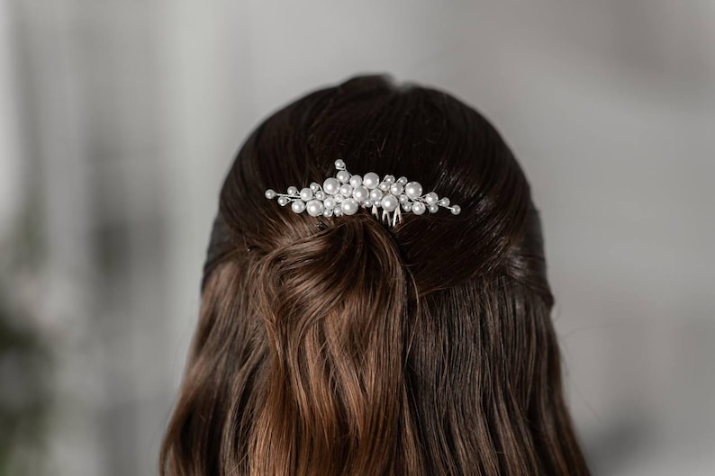 May include: A silver hair comb adorned with clusters of faux pearls. The comb is inserted into dark brown hair, styled in a half-up, half-down hairstyle. The pearls are various sizes and arranged in a floral pattern.