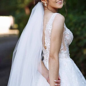 May include: A bride in a white lace wedding dress with a long veil, holding a bouquet of flowers. She is smiling and looking over her shoulder.