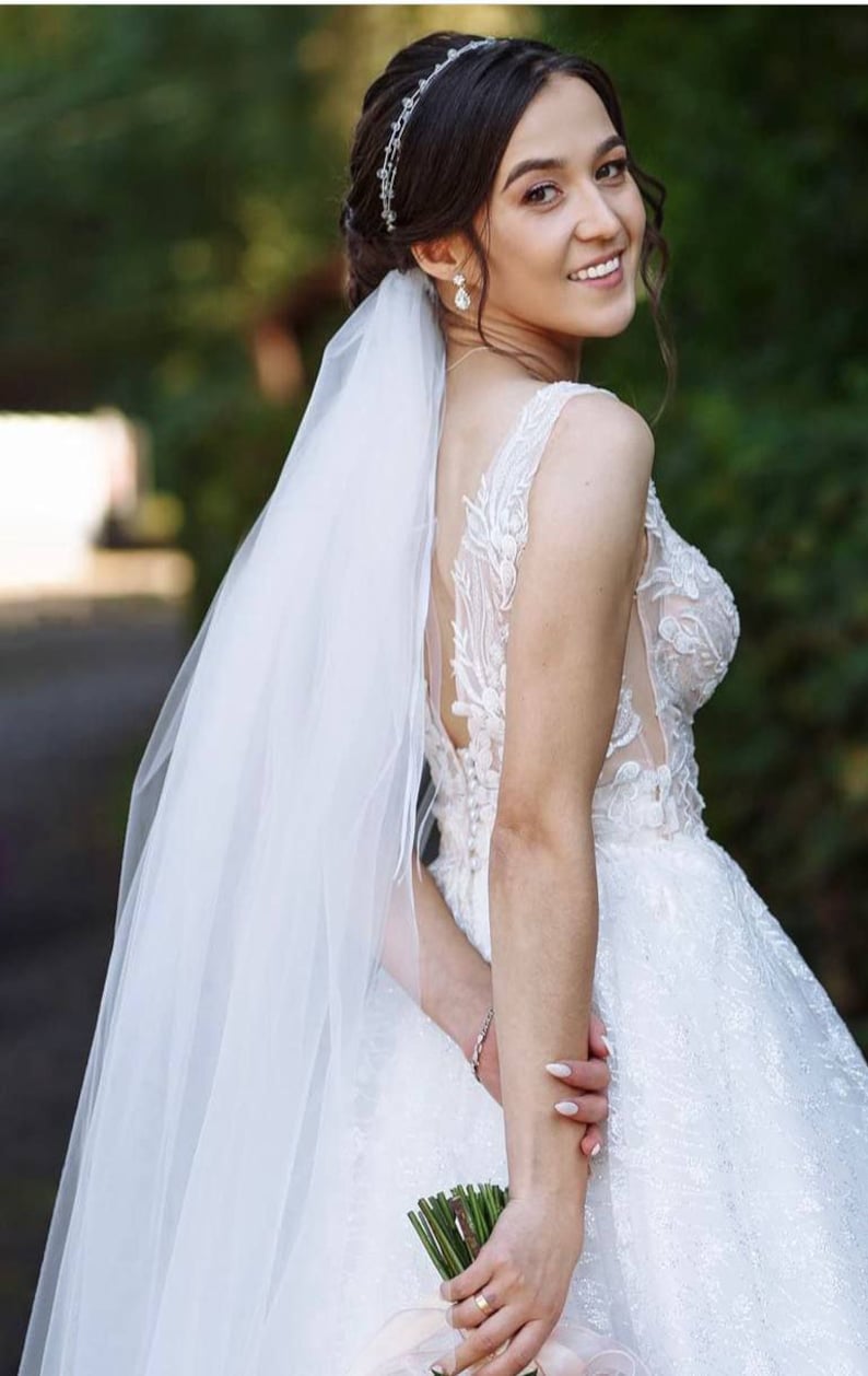 May include: A bride in a white lace wedding dress with a long veil, holding a bouquet of flowers. She is smiling and looking over her shoulder.