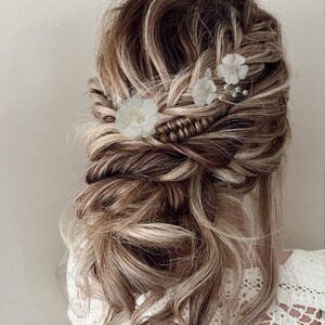 以下が含まれることがあります： 長い茶色の髪を、ゆるいお団子にした編み込みヘアスタイルにした女性。髪には白い花のヘアピンで飾られています。
