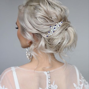 May include: A woman's hairstyle with light blonde, wavy hair, styled in a low bun. A decorative hair comb with white pearls and blue accents is placed in the hair. The woman is wearing a white dress with lace detailing and pearl earrings.
