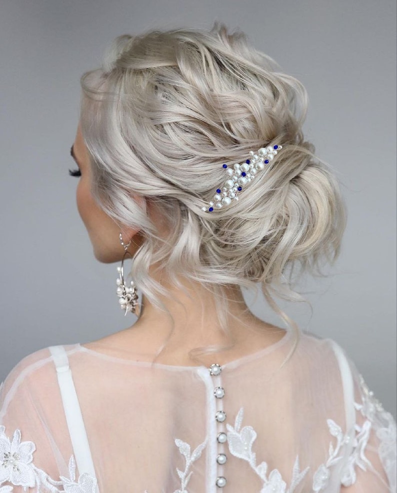 May include: A woman's hairstyle with light blonde, wavy hair, styled in a low bun. A decorative hair comb with white pearls and blue accents is placed in the hair. The woman is wearing a white dress with lace detailing and pearl earrings.
