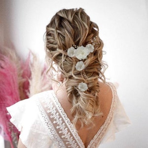 以下が含まれることがあります： 白い花柄のヘアクリップでゆるく編まれたブロンドの髪をした女性。レースのディテールが施された白いドレスを着ています。