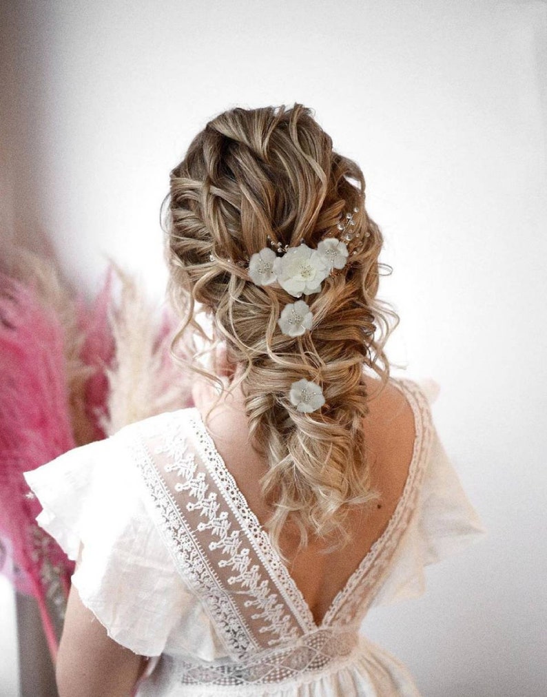 以下が含まれることがあります： 白い花柄のヘアクリップでゆるく編まれたブロンドの髪をした女性。レースのディテールが施された白いドレスを着ています。