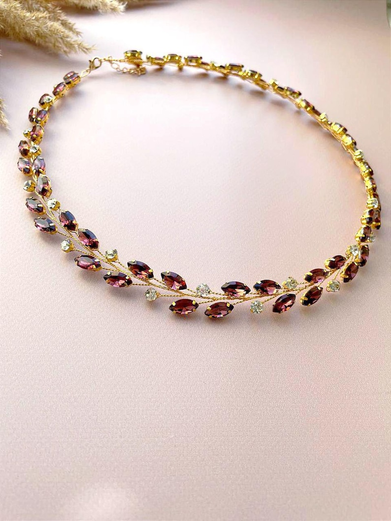 May include: A delicate gold necklace with a double row of marquise-shaped purple rhinestones and small clear rhinestones. The necklace is designed to resemble a vine with leaves.