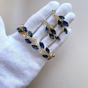 May include: A pair of gold earrings with dangling blue and clear crystal stones. The earrings are shaped like a branch with leaves.