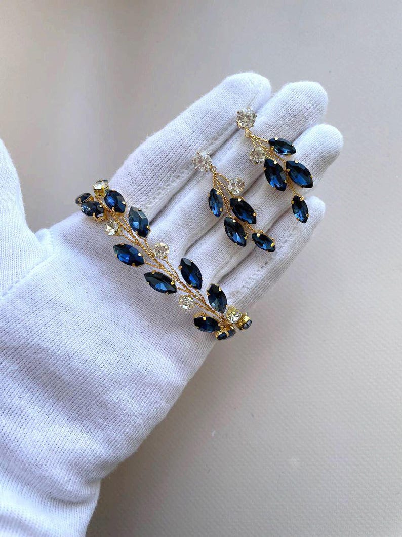 May include: A pair of gold earrings with dangling blue and clear crystal stones. The earrings are shaped like a branch with leaves.