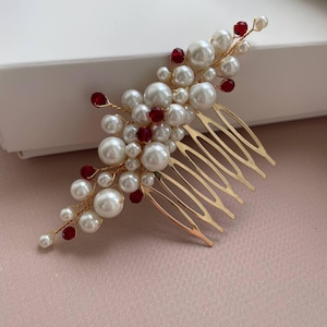 May include: A gold-toned hair comb adorned with clusters of faux pearls and red beads. The comb features a series of tines for securing in hair. The pearls vary in size, creating a textured, elegant look. The red beads add a pop of color.